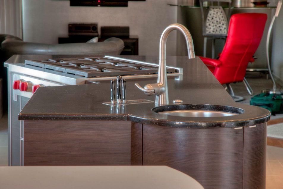 Modern kitchen island with stainless sink, faucet, and dark wood cabinets; red chair in the background