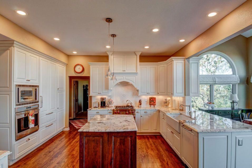 Bright white kitchen with island, wood floors, built-in ovens, and large window overlooking greenery
