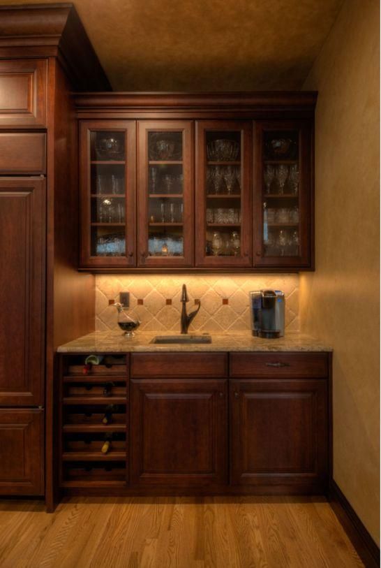 Warmly lit built-in kitchen bar with dark wood cabinets, glass-front uppers, and a stone countertop.