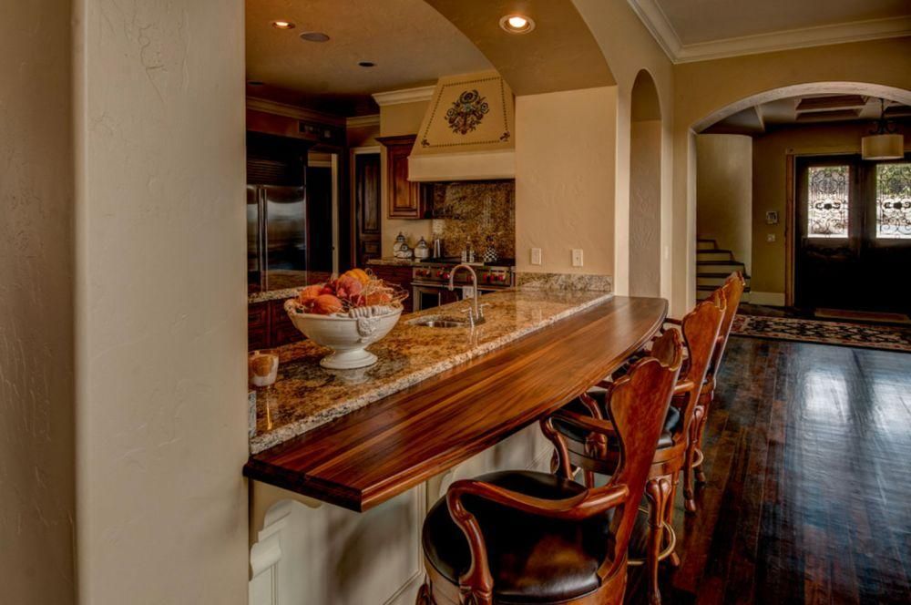 Elegant home bar with granite counter, wooden stools, and dark hardwood floors opening to a living room.