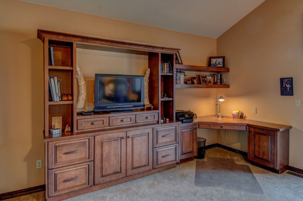 Home office corner with wooden built-in desk, TV cabinet, shelves, lamp, and beige walls.