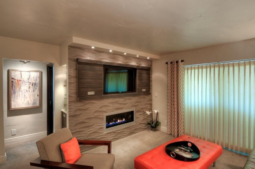 Modern living room with wall-mounted TV, fireplace, beige seating, and orange ottoman near vertical blinds.