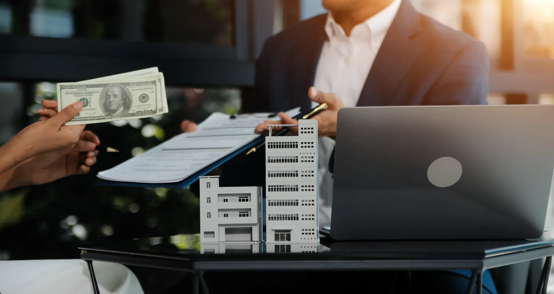 Person handing cash to someone in a suit, with a building model, documents, and laptop on the table.