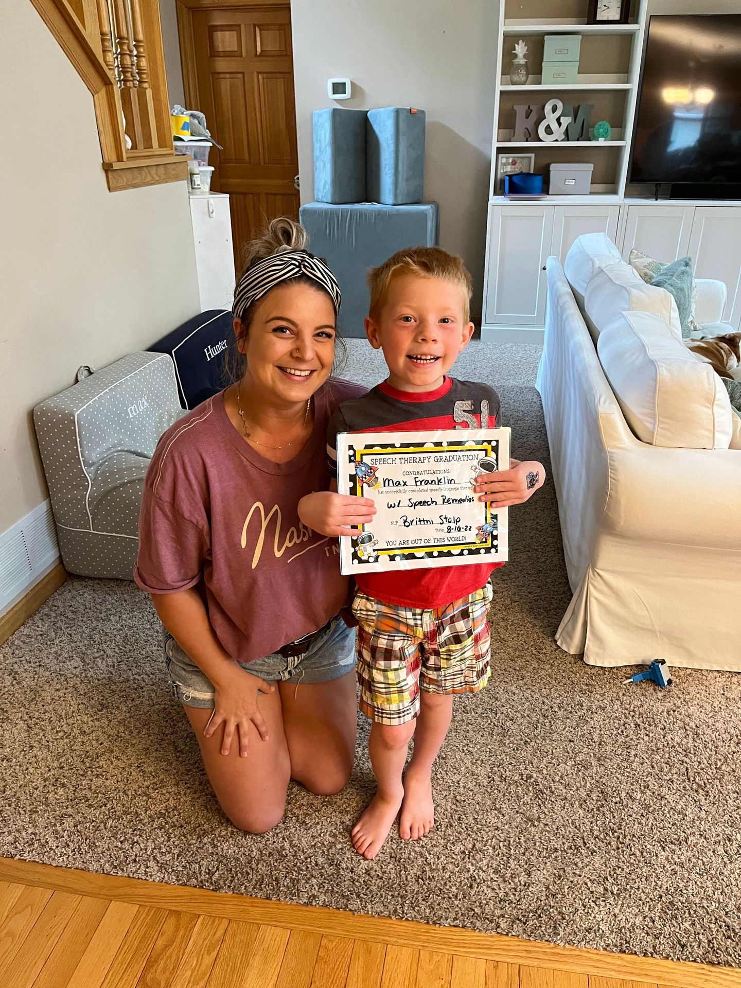 A woman is kneeling next to a young boy holding a certificate in a living room.
