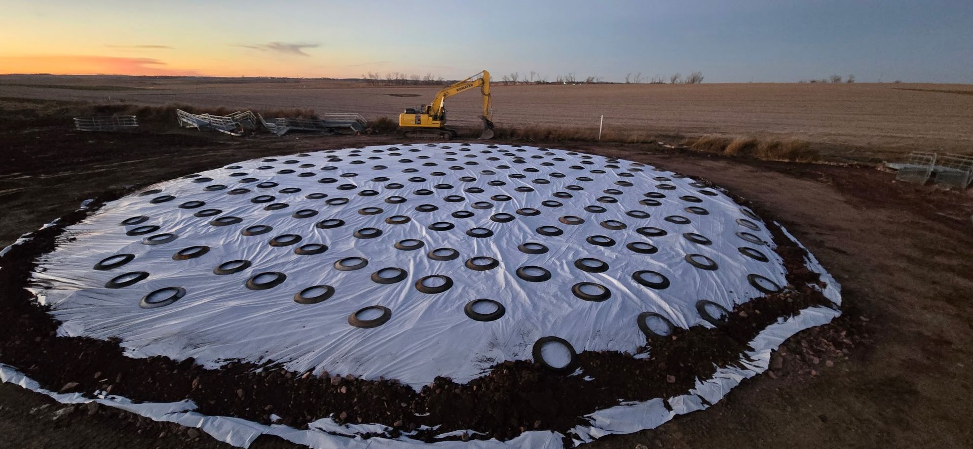 A wide-angle aerial view of a construction site featuring a white circular tarp weighed down by evenly spaced tires.