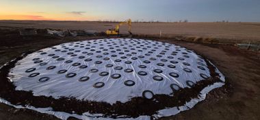 A wide-angle aerial view of a construction site featuring a white circular tarp weighed down by evenly spaced tires.