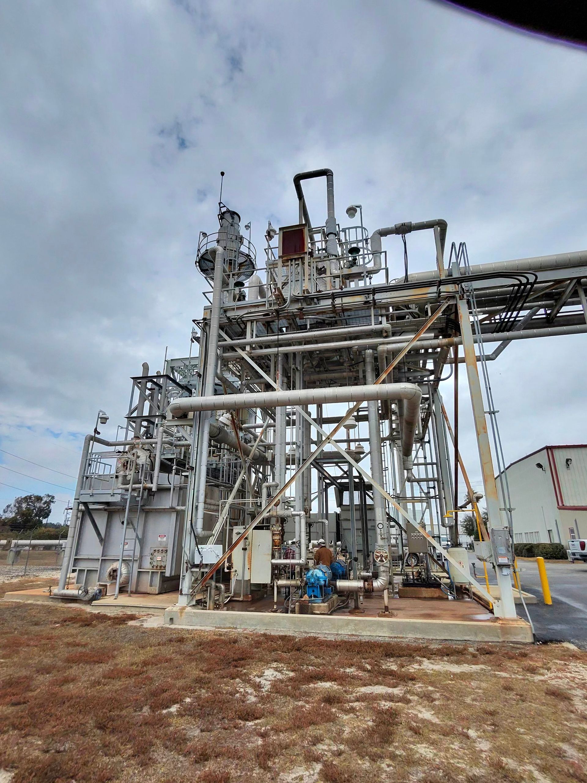 Industrial refinery equipment with silver pipes and a worker in blue helmet under a cloudy sky