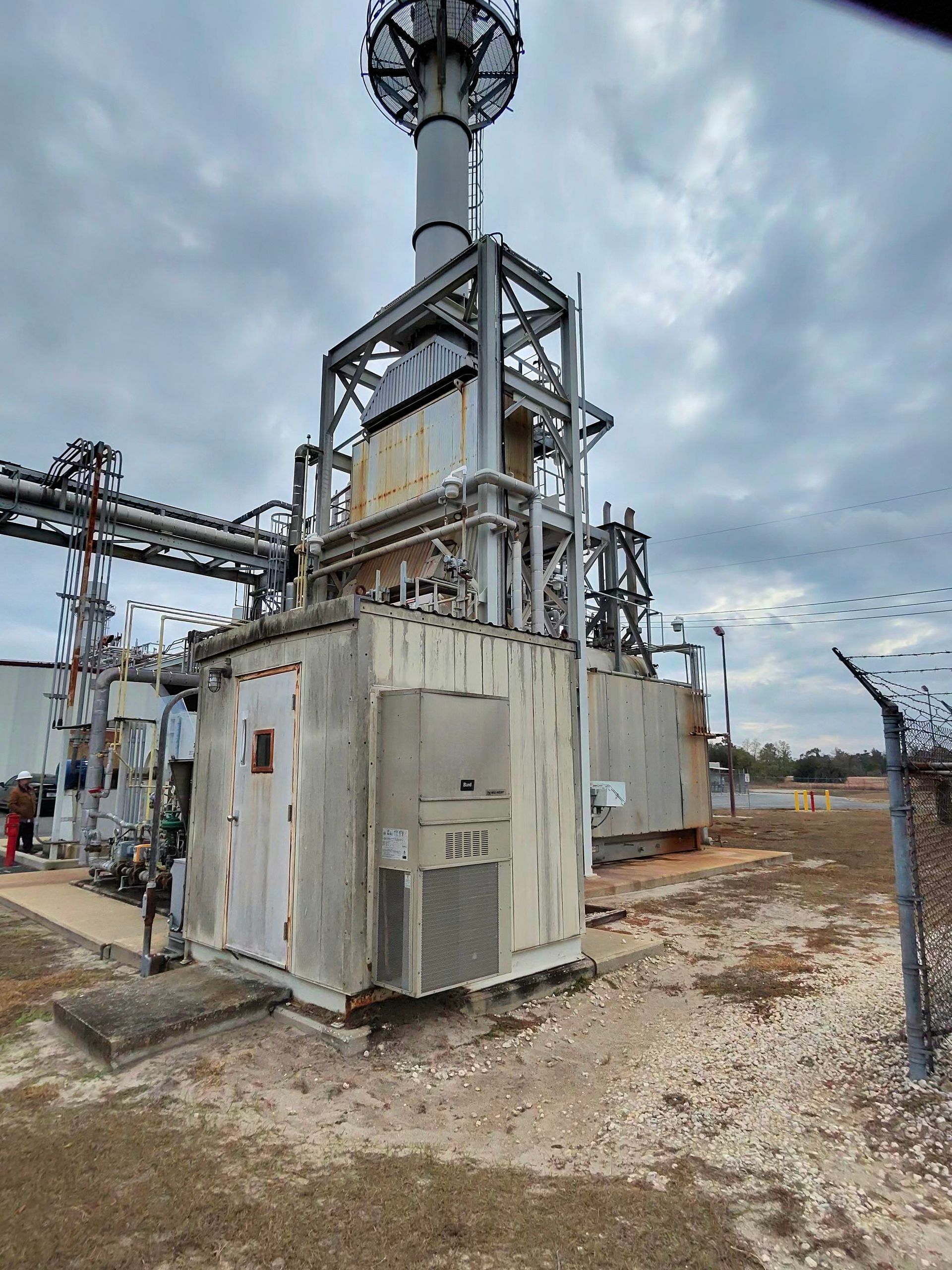 Industrial facility with metal piping, a beige equipment container, and a tall steel tower on a gravel lot