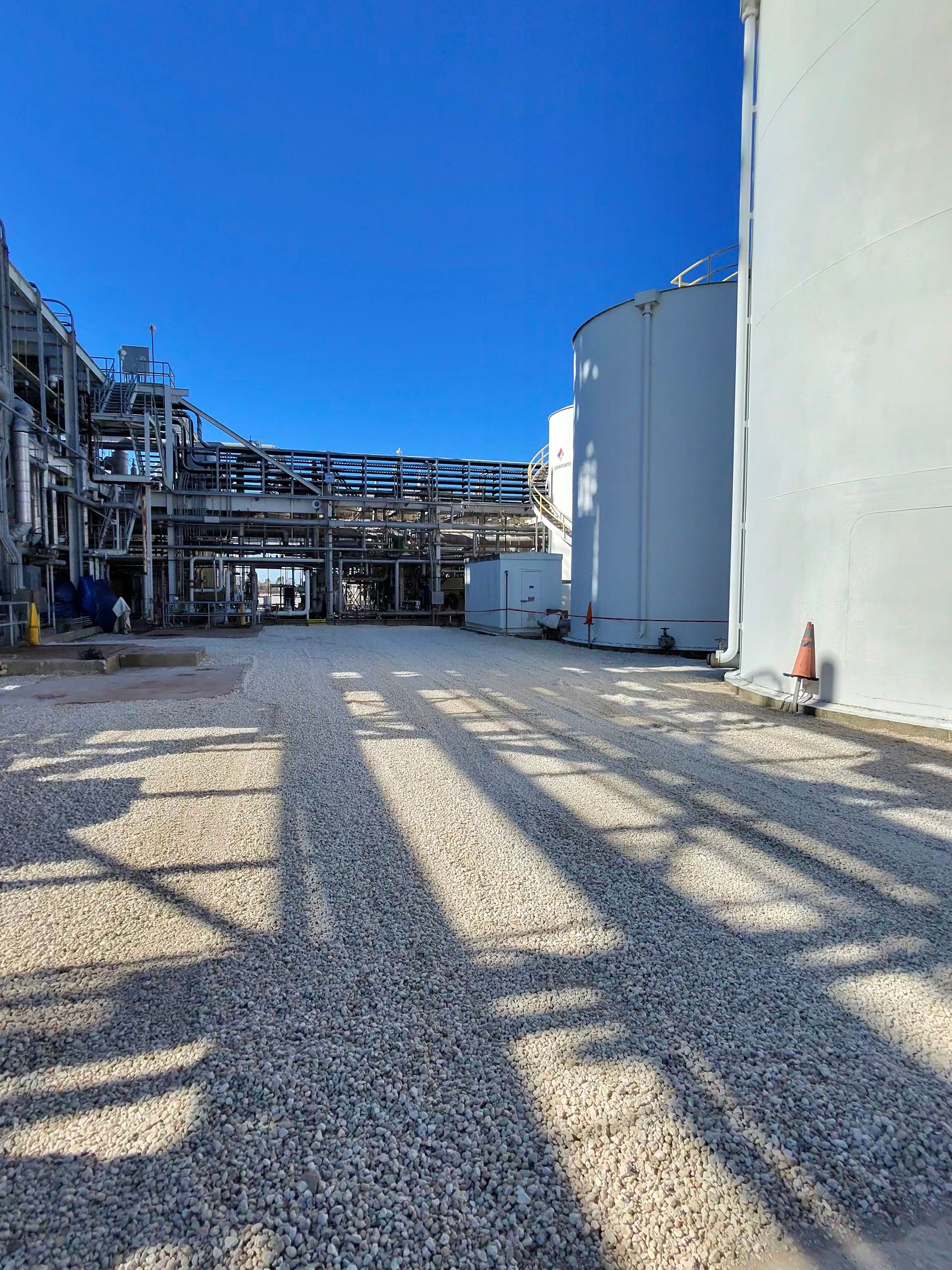 Industrial plant yard with gravel ground, large silver tanks, piping, and strong shadows under a blue sky