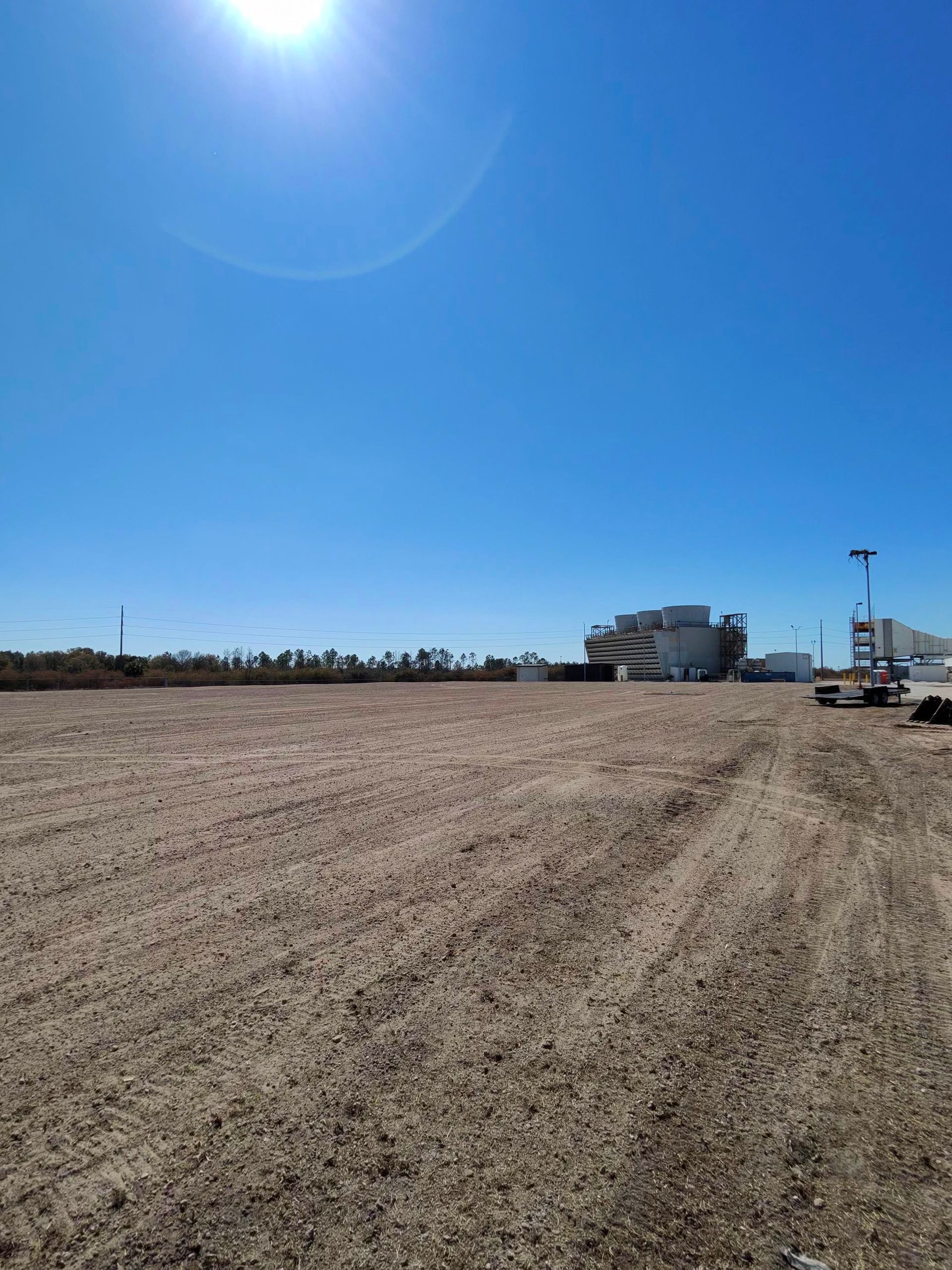 Wide empty dirt lot under a bright blue sky, with a few buildings and vehicles in the distance.