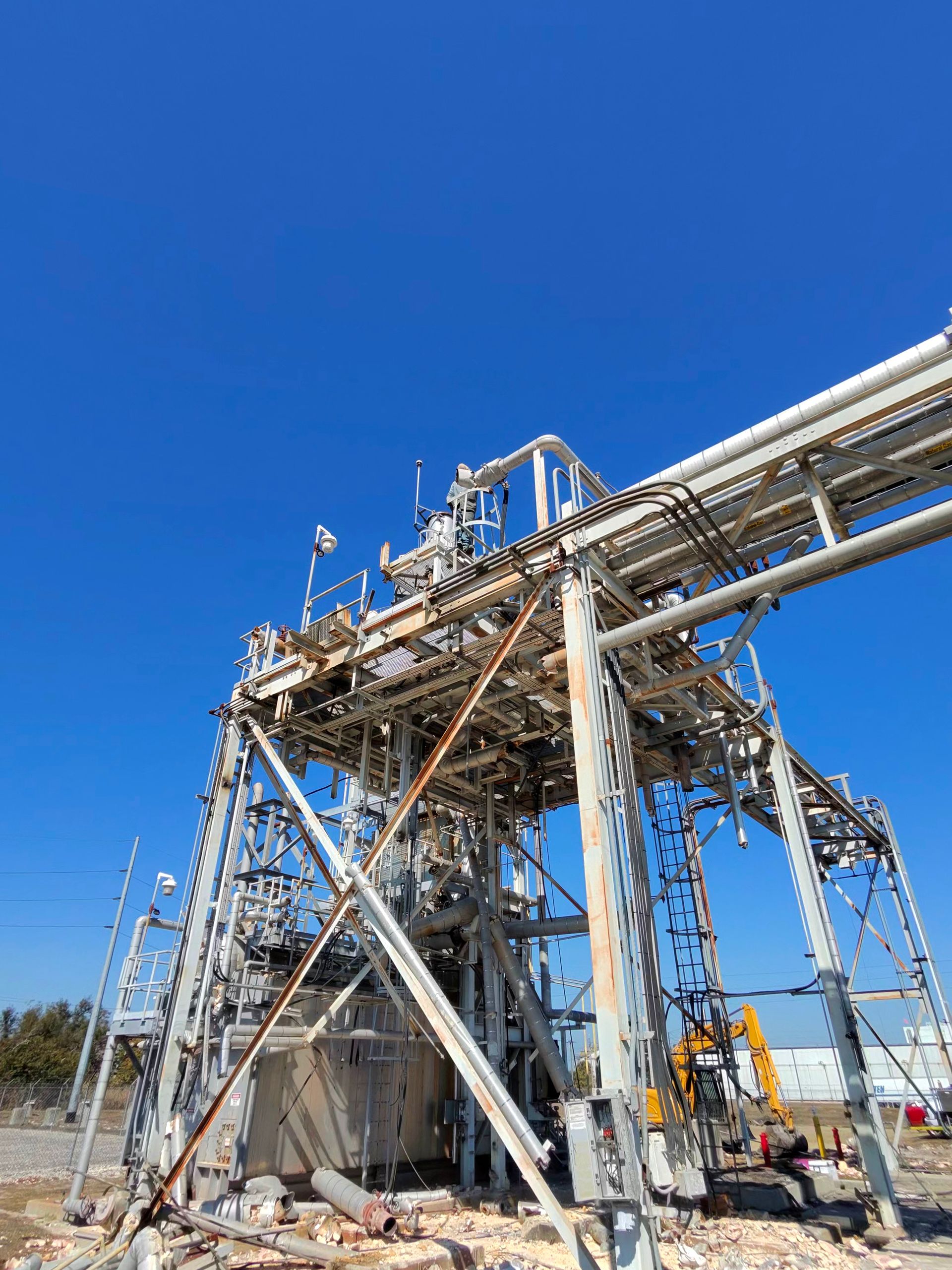 Industrial steel structure at a plant under a clear blue sky, with pipes and scaffolding.