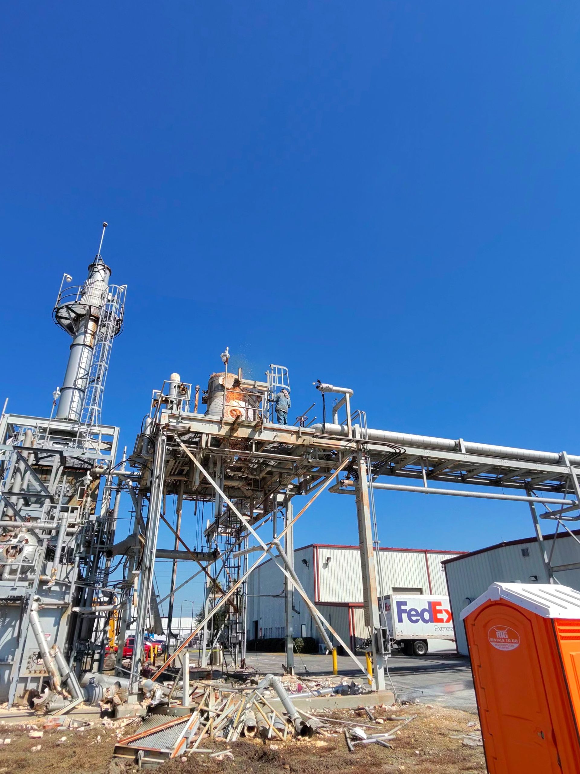 Industrial facility with metal piping and tower under a clear blue sky, orange portable toilet in foreground