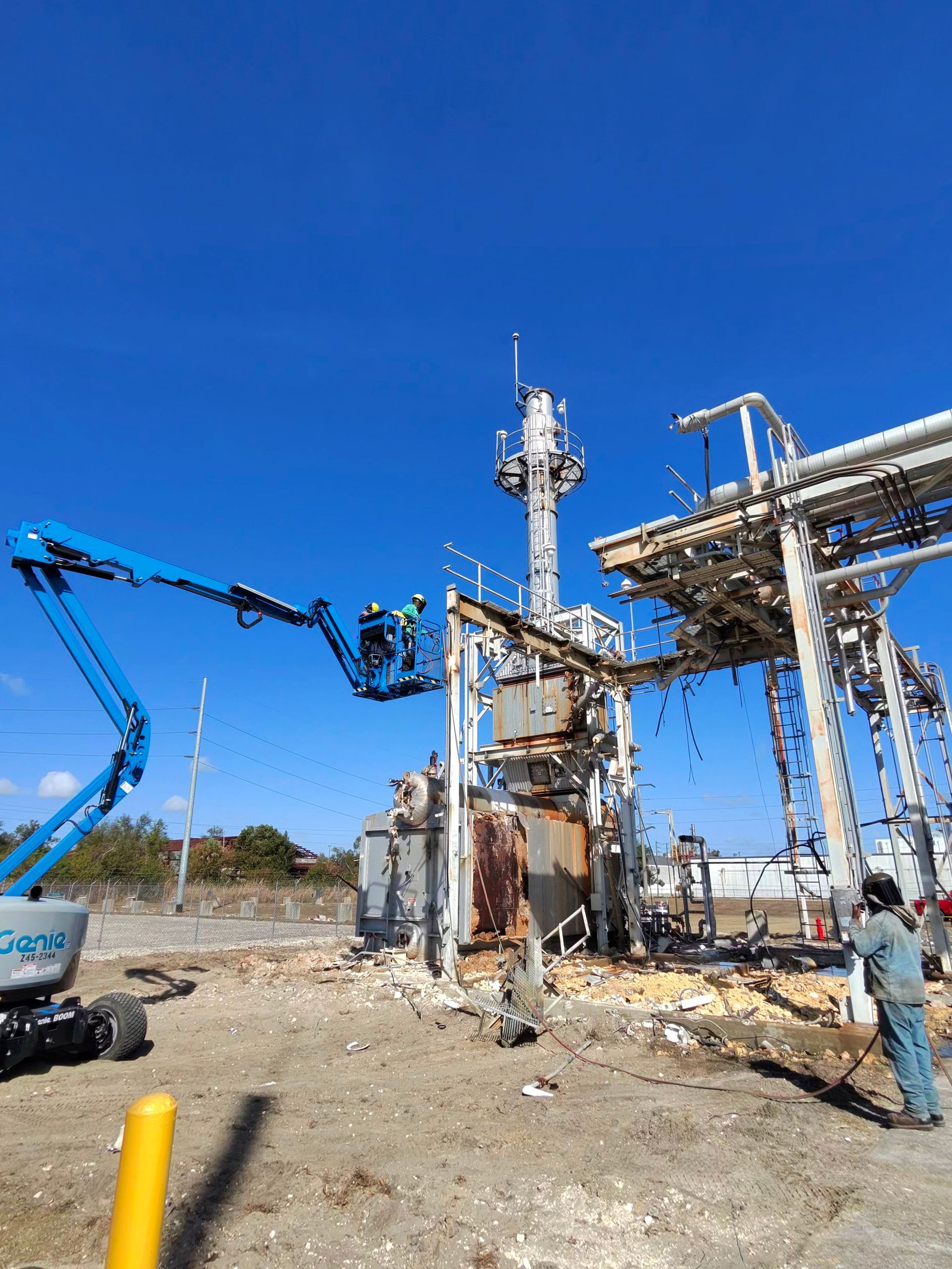 Industrial site with blue lift and workers around piping and metal structures under a clear sky