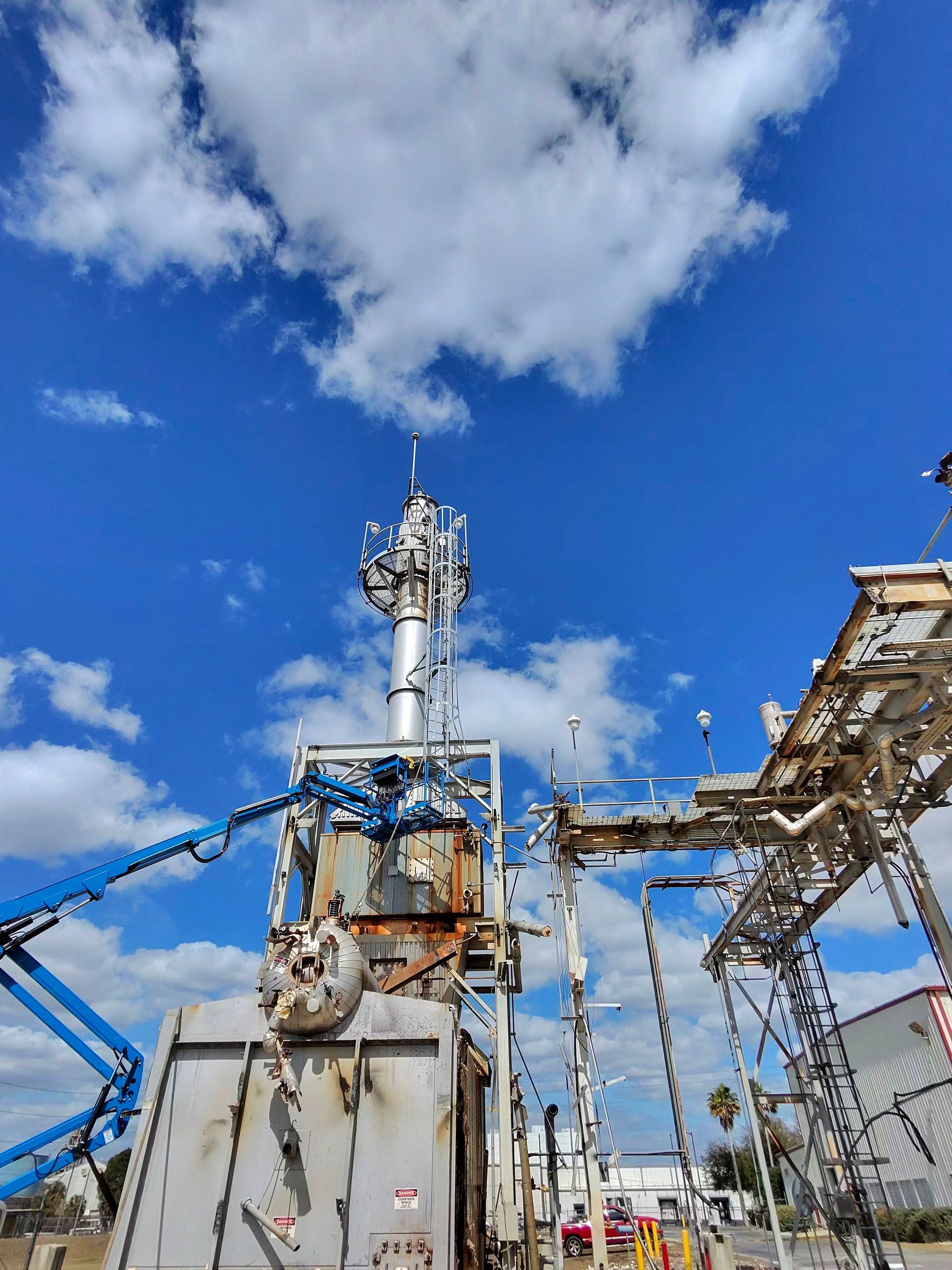 Industrial oilfield machinery under a blue sky with clouds, including pipes, a tank, and metal structures