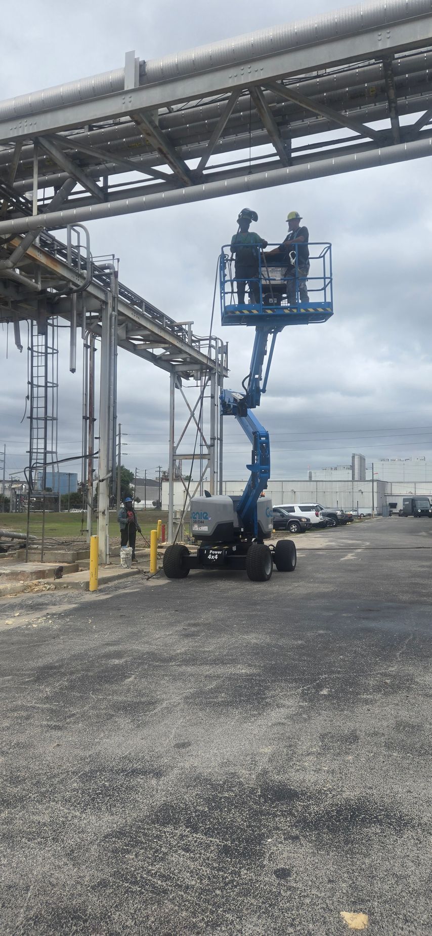 Two workers in a blue boom lift beneath industrial piping in a refinery yard