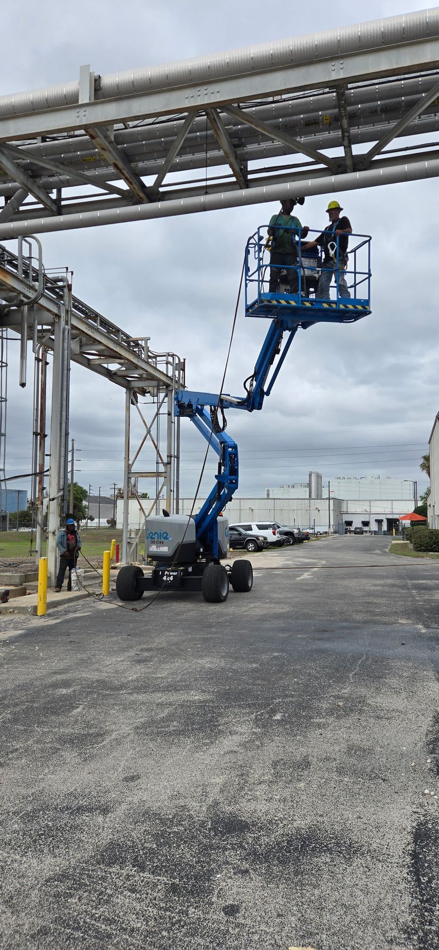 Blue boom lift raising two workers beside industrial piping under an overcast sky