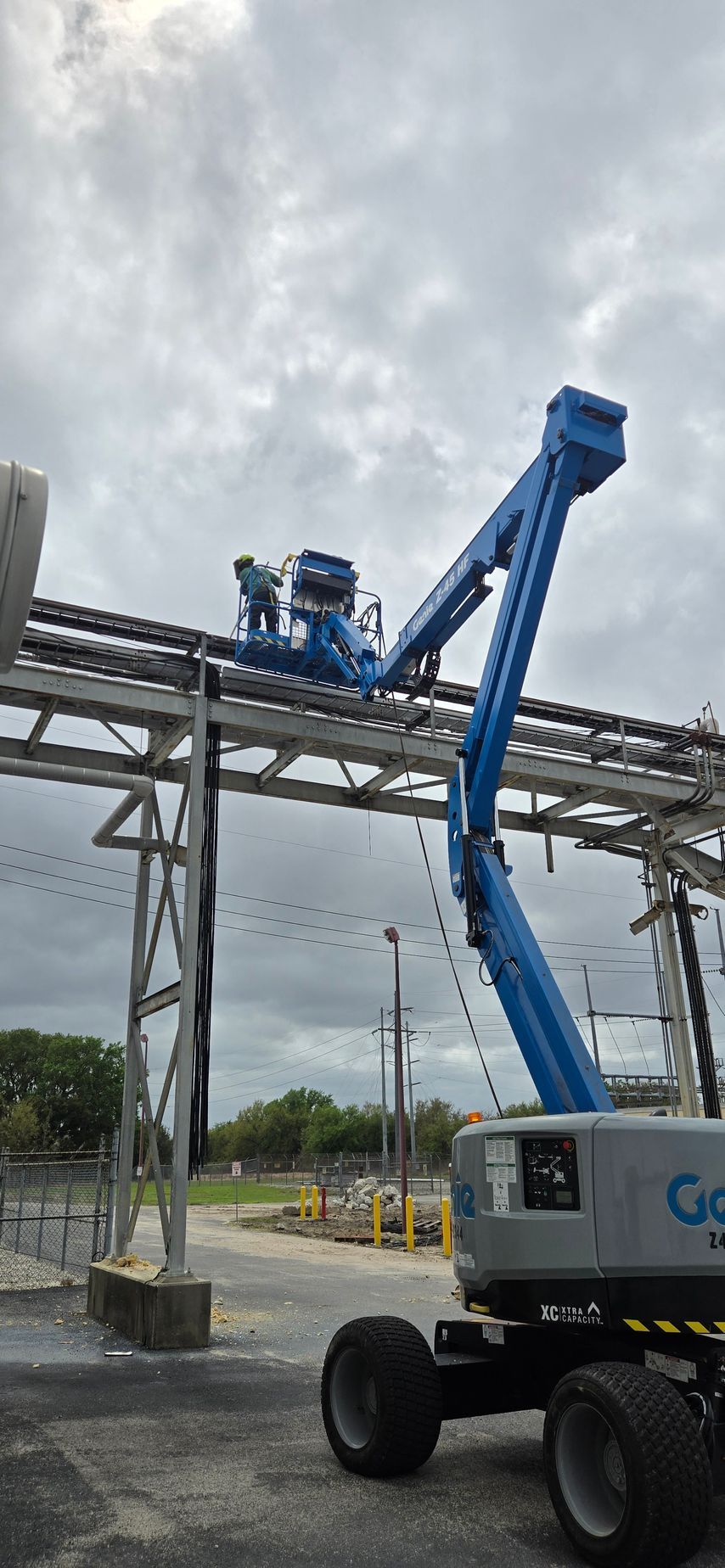 Blue boom lift operating beside an elevated conveyor structure at an industrial site