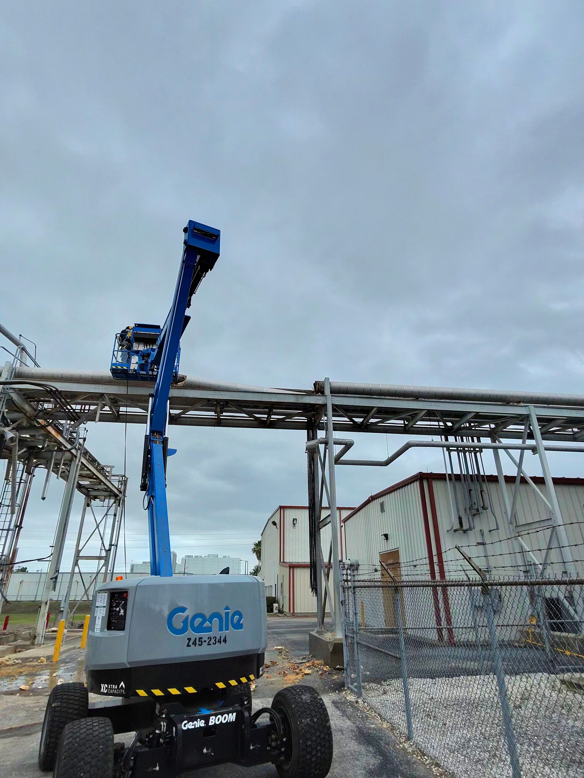 Blue Genie boom lift raised beside industrial piping at a fenced work site under a cloudy sky