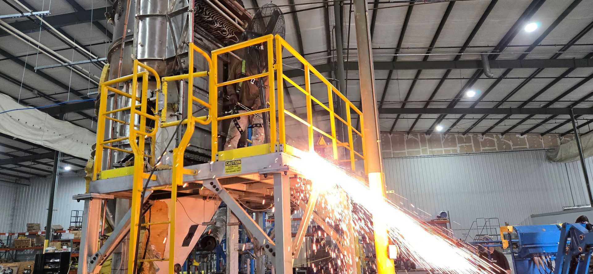 Industrial machinery ejecting sparks in a factory, with a yellow metal platform overhead.