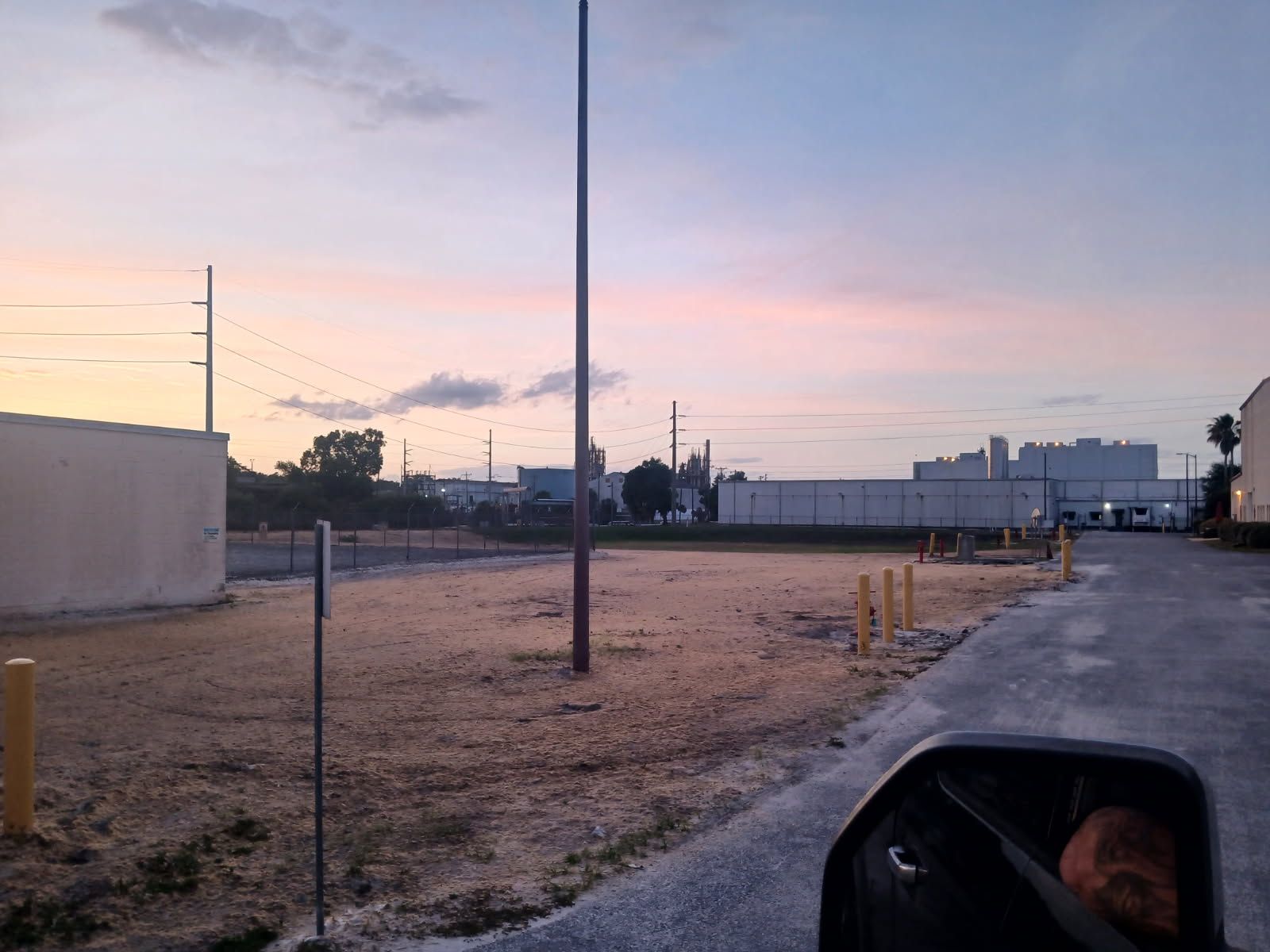 Dirt lot at dusk with utility poles, parked trucks, and a side mirror showing a woman’s face.