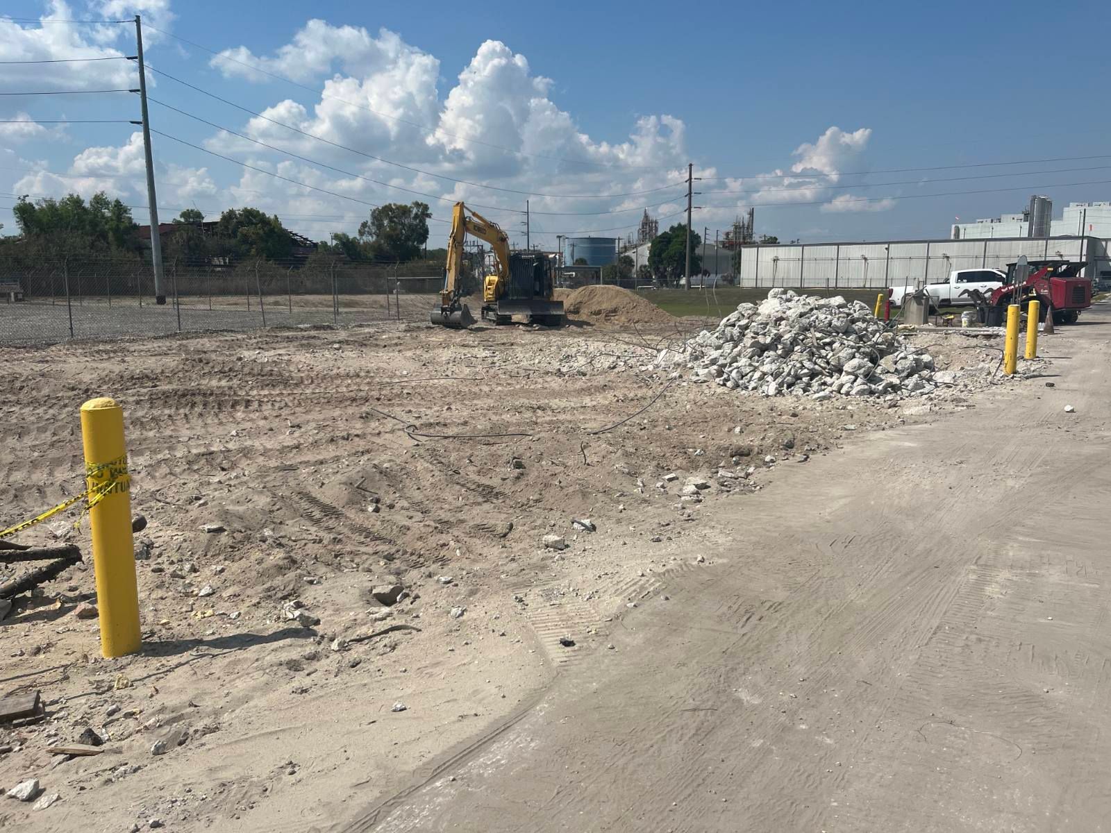 Construction site with excavator, dirt lot, and piles of rubble beside a paved road