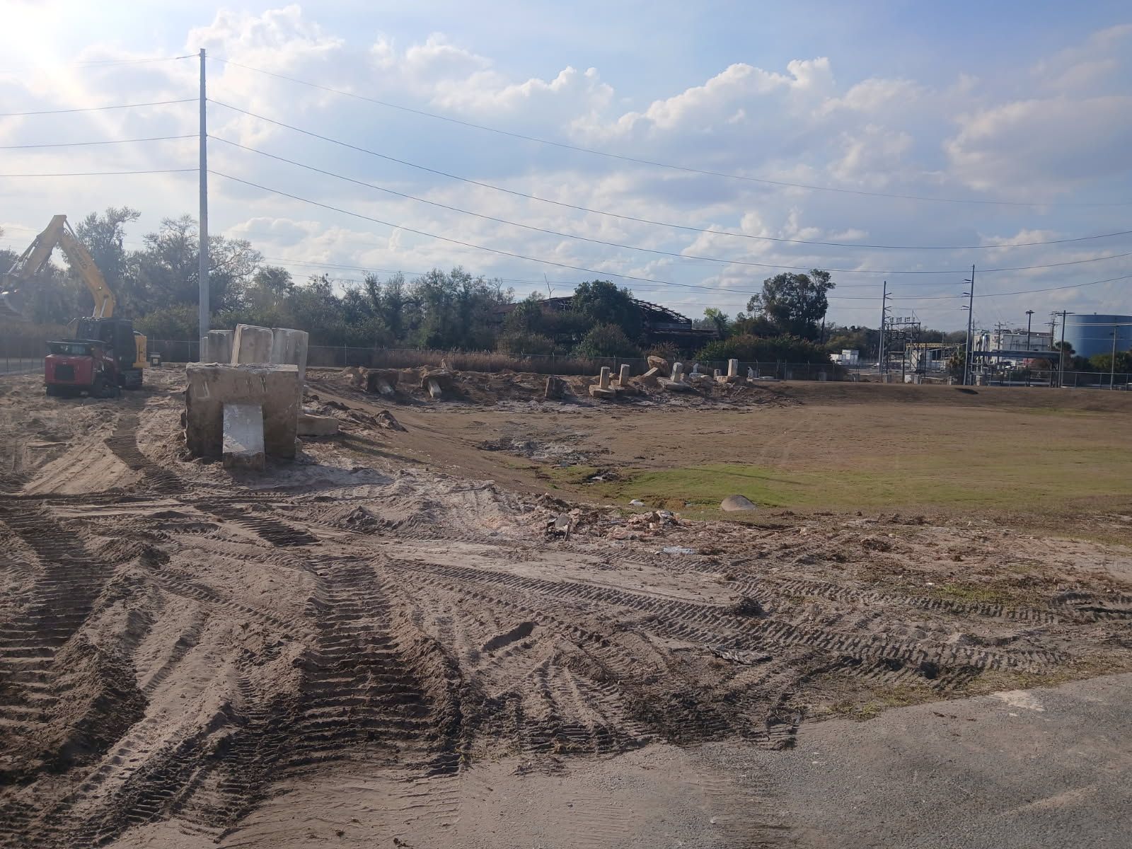 Muddy construction site with tire tracks, excavator, and utility poles under a cloudy sky