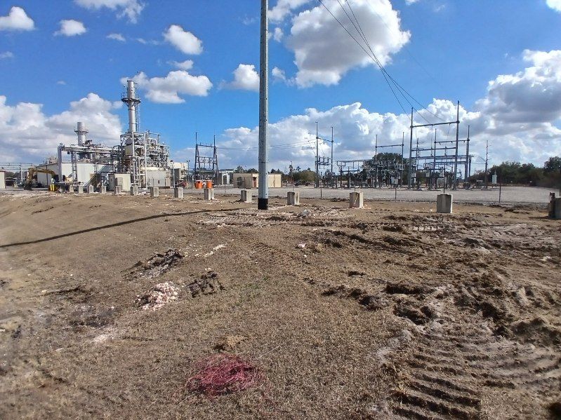 Industrial plant under a cloudy blue sky, with dirt foreground and power lines in the distance