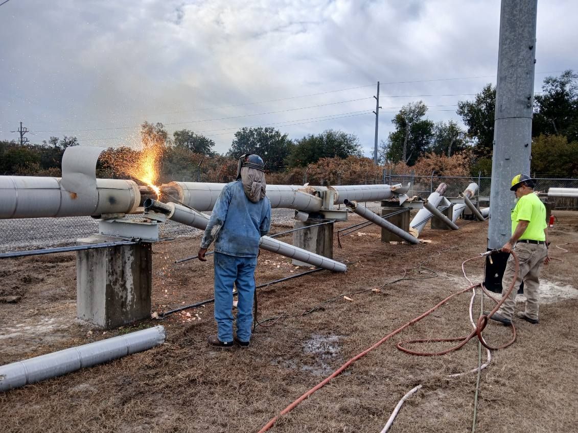 Utility workers inspect a damaged power line as flames flare near fallen poles on a muddy roadside.