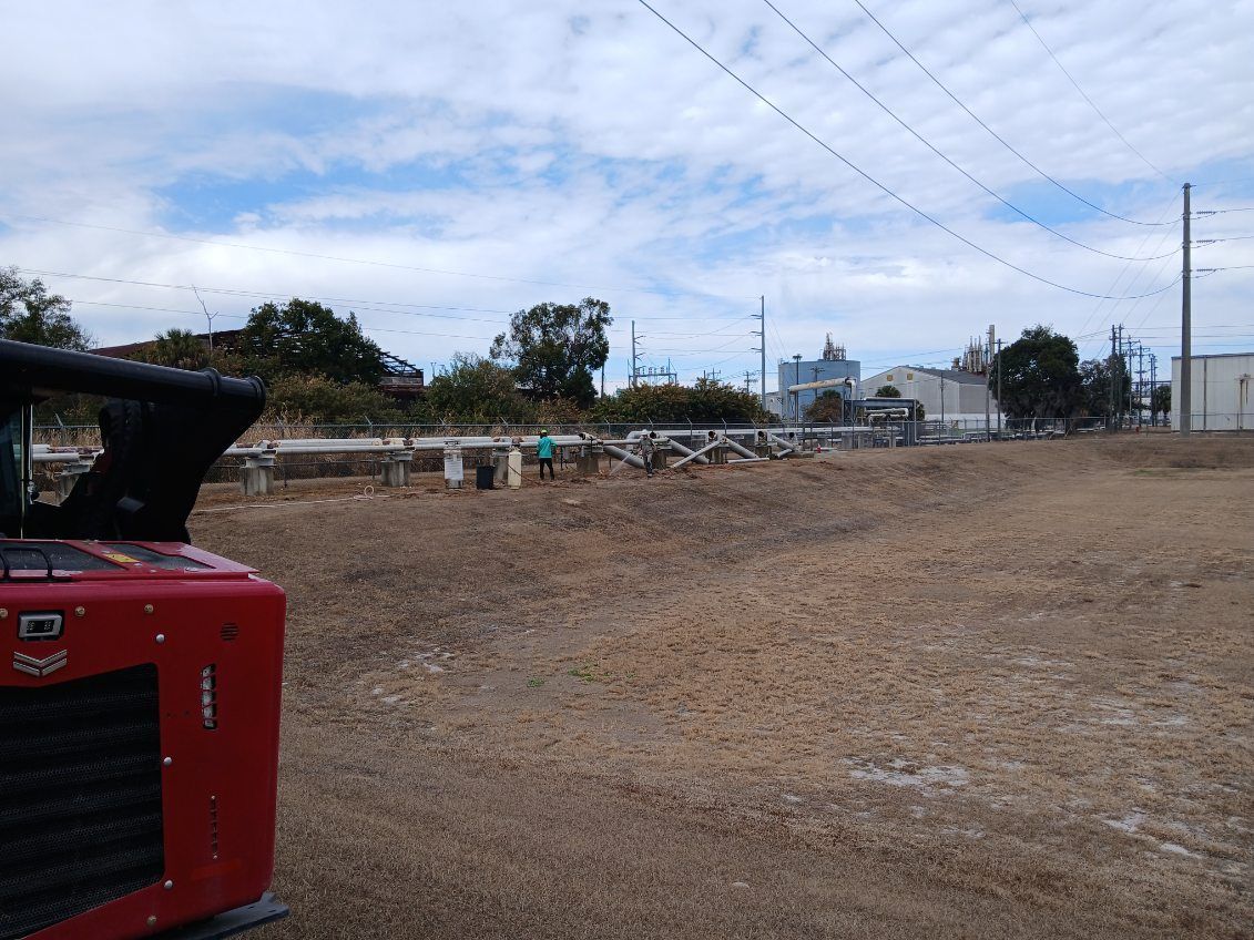 Open dirt lot with a red tractor at left, under a cloudy sky, near a fence and small buildings.