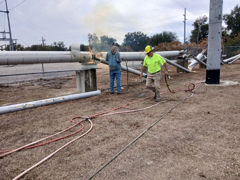 Two workers in safety gear inspect a gas line near a pipeline, with smoke in the background.
