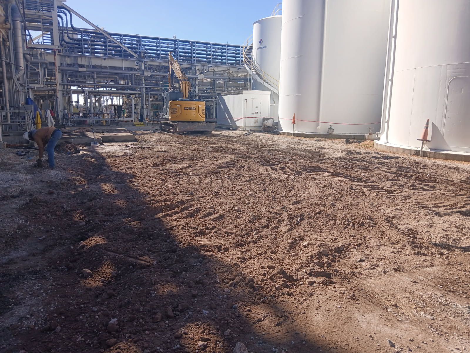 Industrial site with large white storage tanks, steel piping, a yellow loader, and muddy ground under construction