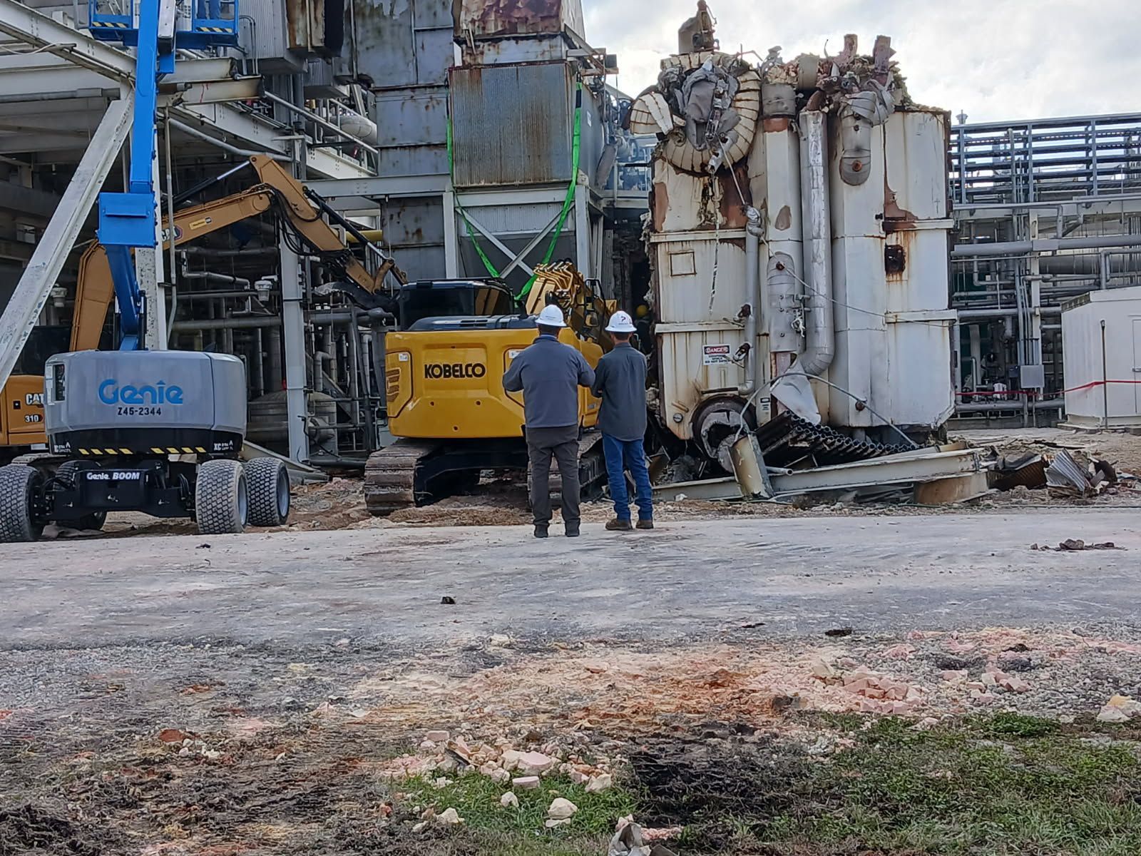 Demolition site with two workers in hard hats and a yellow excavator among rubble and damaged industrial buildings