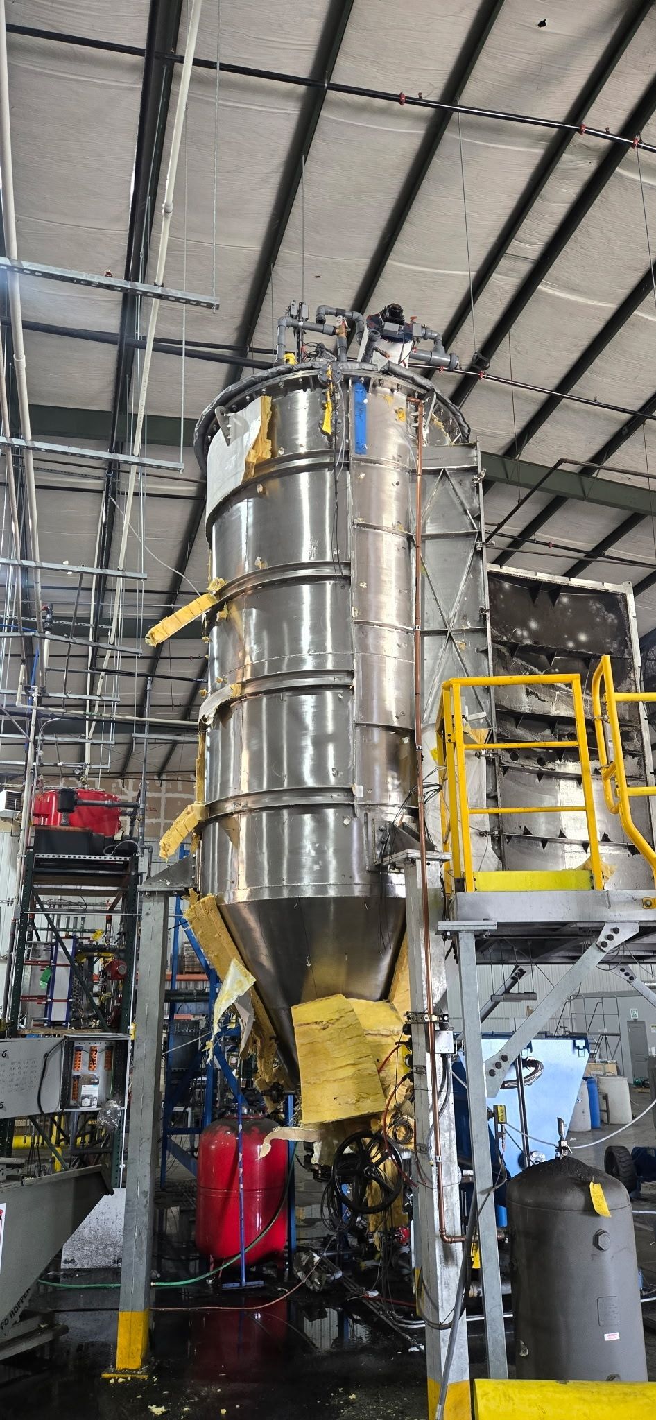 Stainless steel industrial hopper with yellow piping in a factory setting