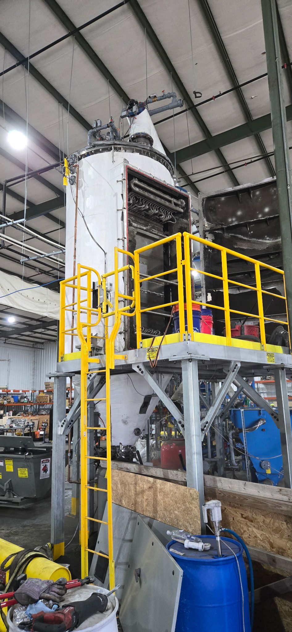 Industrial silo and yellow-railed platform inside a factory, with ladder and machinery below.