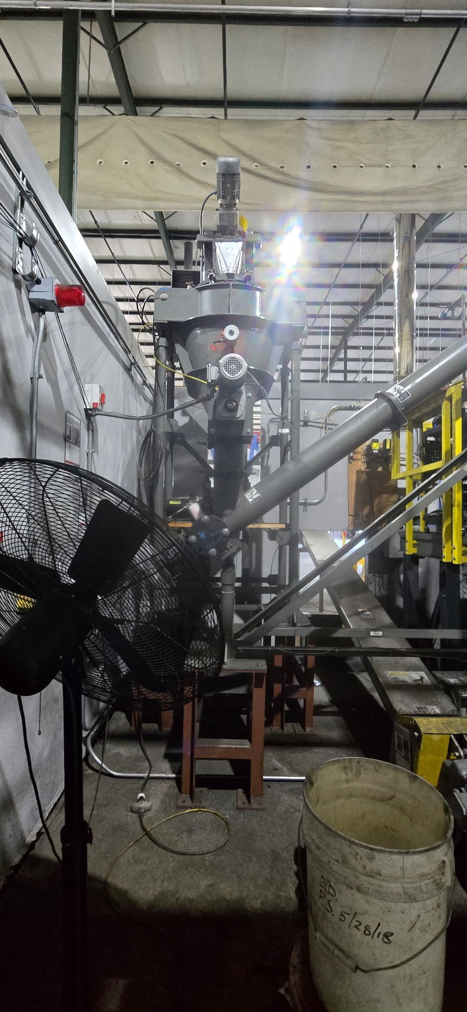 Industrial facility with a large metal mixer and pipes, lit by a bright overhead light, with a fan and bucket in front.