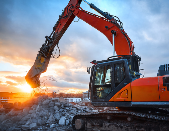 An orange excavator with a hydraulic attachment works on a pile of concrete debris against a golden sunset sky.