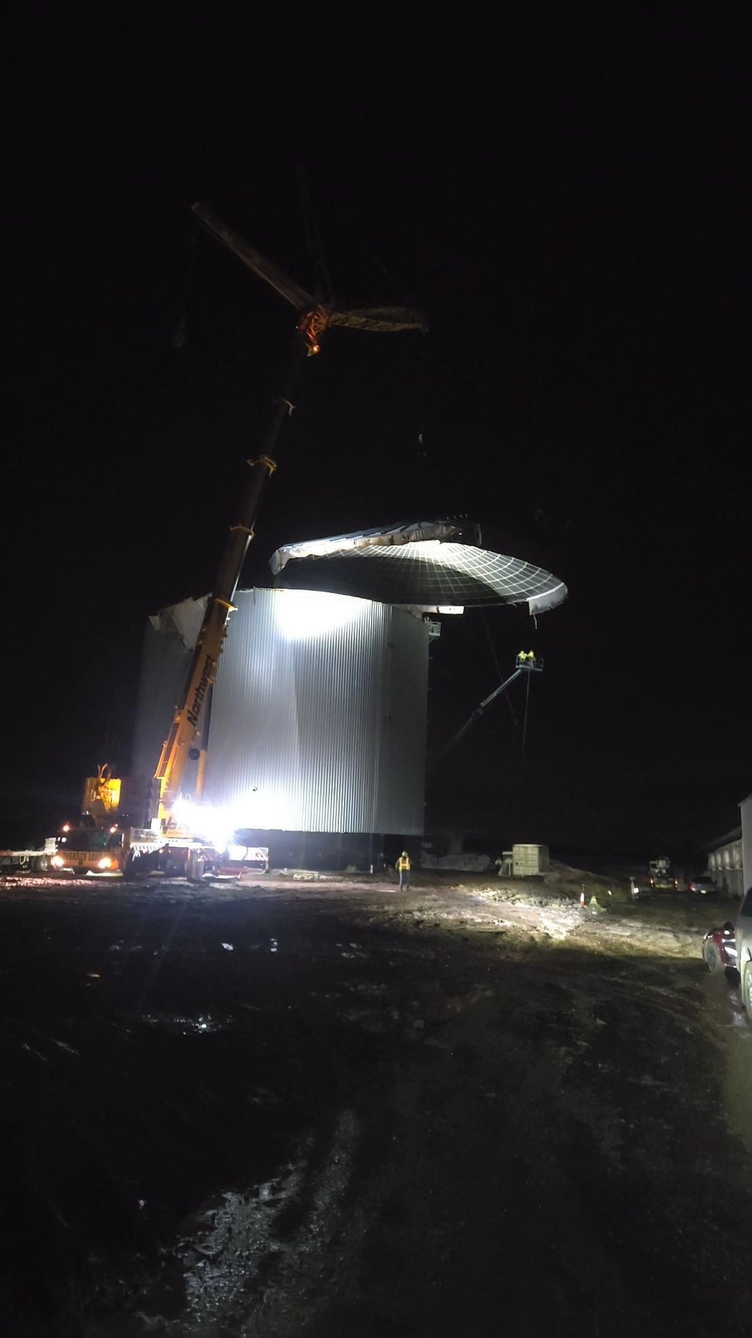 Night construction site with a crane lifting a large illuminated curved structure over wet ground