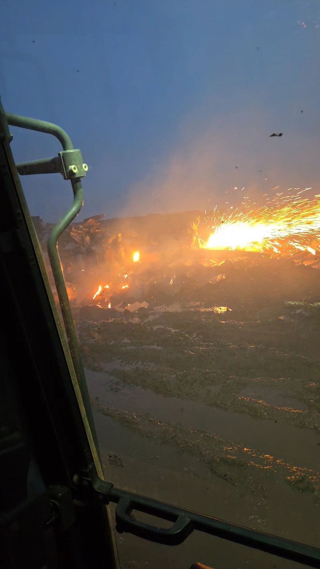 View from a vehicle cabin of sparks flying over a dark, muddy field at dusk.