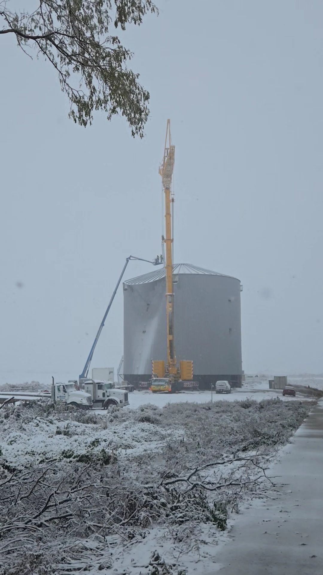 A crane and a cherry picker work on a large, silver storage tank in a snowy, winter landscape with downed tree branches.