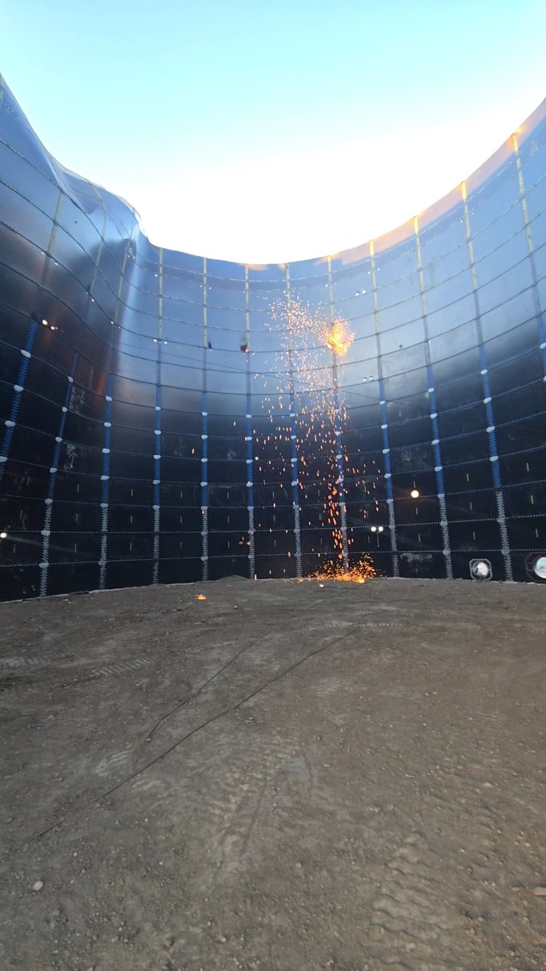 Sparks fly from welding work on the interior metal wall of a large, circular storage tank under construction.