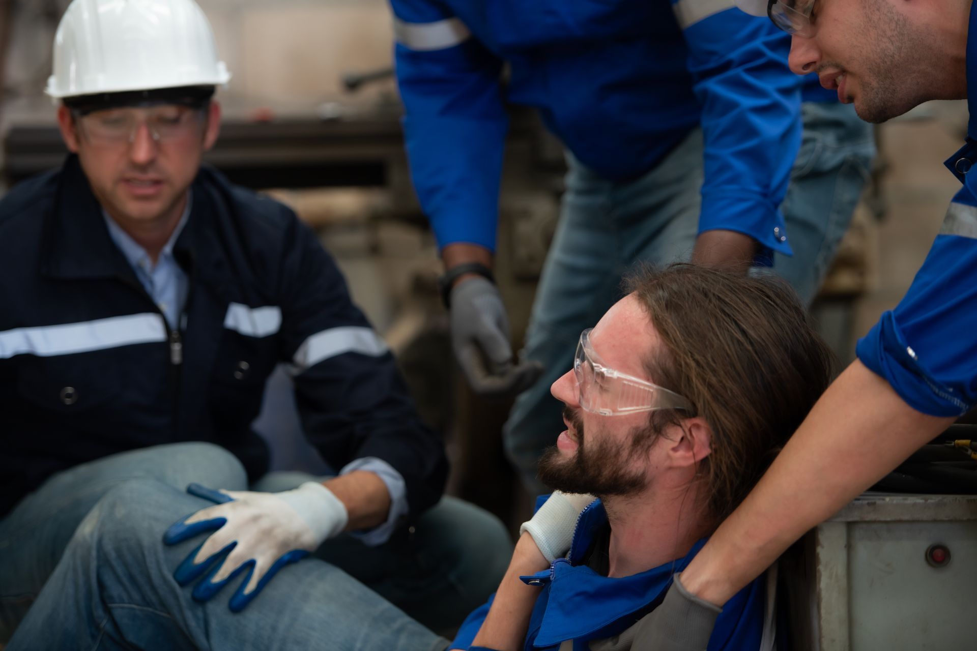 Workers assisting an injured colleague on a factory floor. One man is sitting on the floor, in pain.