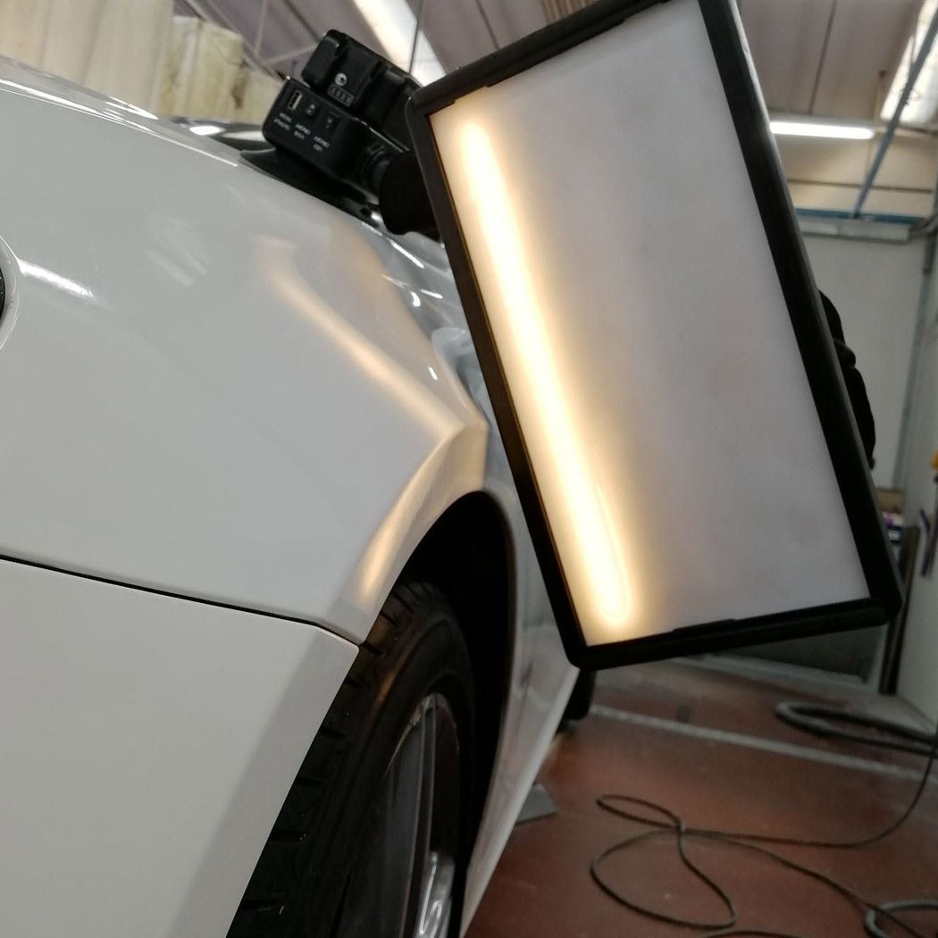 Un'auto bianca viene ispezionata in un garage, con un grande pannello luminoso rettangolare accanto alla ruota anteriore.