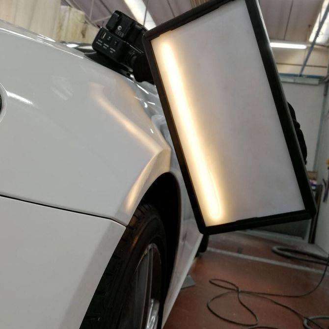 Un'auto bianca in un'officina con un pannello rettangolare illuminato fissato nella zona della ruota anteriore.