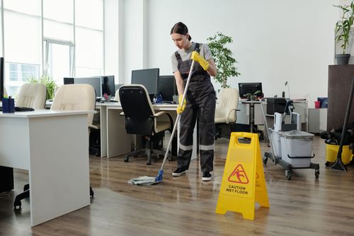 A person in gray work coveralls mops a modern office floor near a yellow 