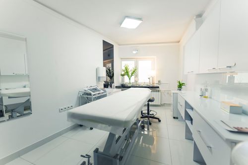 A bright, minimalist medical or aesthetic treatment room featuring a white patient examination table and storage cabinets.