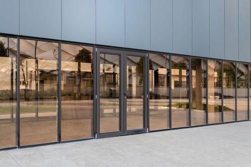 A glass-paneled building exterior featuring a set of double entry doors framed in dark metal against a light concrete floor.