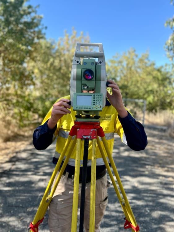 A Man In A Yellow Vest Is Using A Total Station On A Tripod — Point to Point Surveying In Tatura, VIC