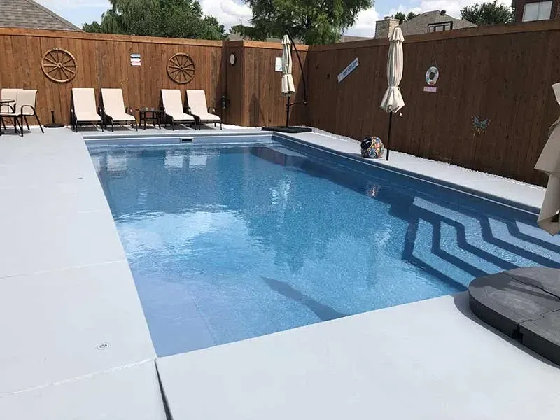 Backyard swimming pool with blue water, lounge chairs, wooden fence, and closed umbrella