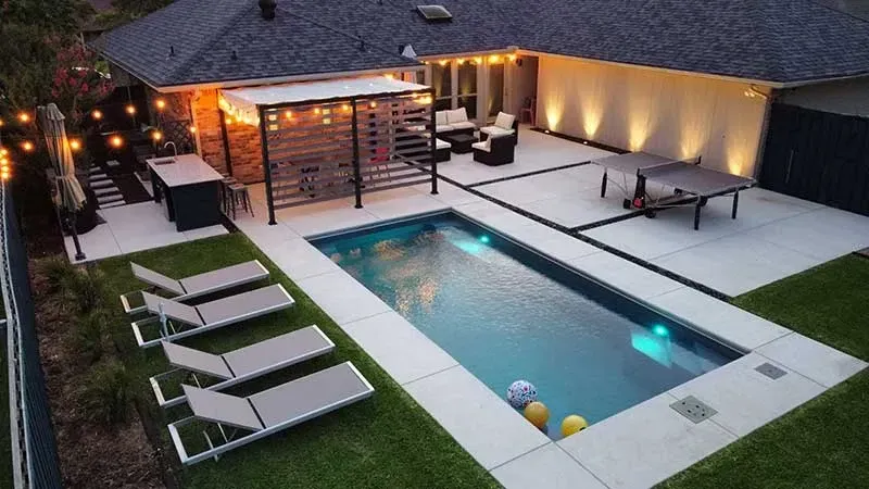 Modern backyard with a narrow lit pool, lounge chairs, patio dining set, and covered outdoor seating area at dusk