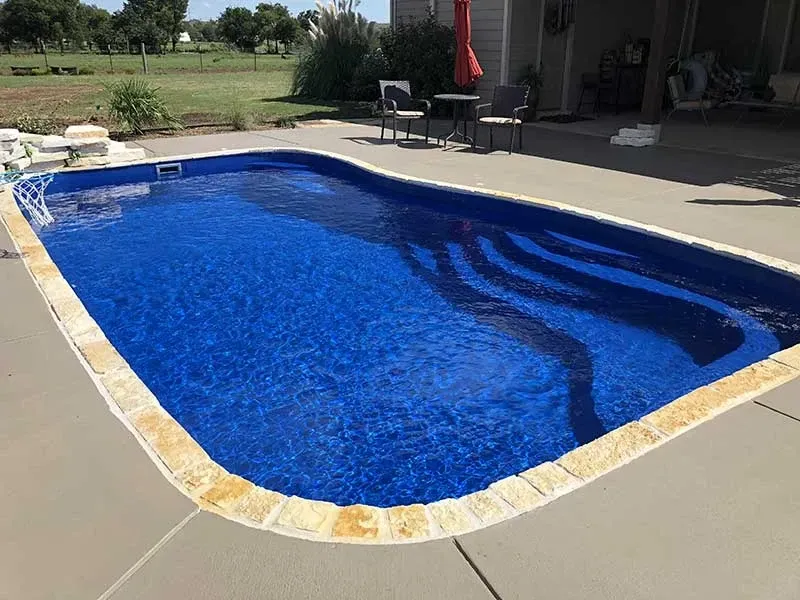 Backyard swimming pool with blue water and tan stone deck in bright sunlight